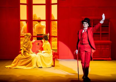 2 actors in yellow costumes in the background, and 1 actor in a red costume in the foreground in the importance of being earnest.